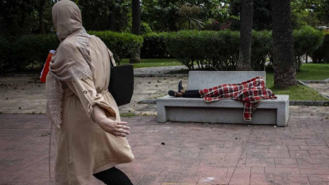 Una mujer observa a Mohamed, un menor marroquí de 17 años que duerme en el banco de los Jardines de Argentina de Ceuta tras entrar a nado a la ciudad a principios de esta semana.- JAIRO VARGAS