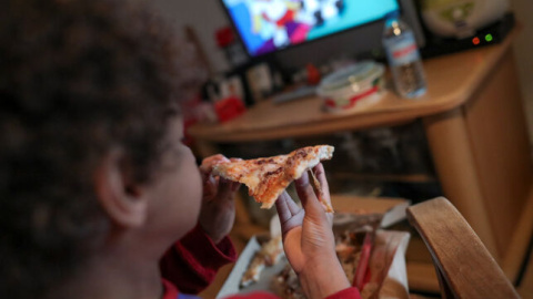 Un niño come un trozo de pizza del menú infantil de Telepizza mientras ve la televisión en su casa. EUROPA PRESS / JESÚS HELLÍN