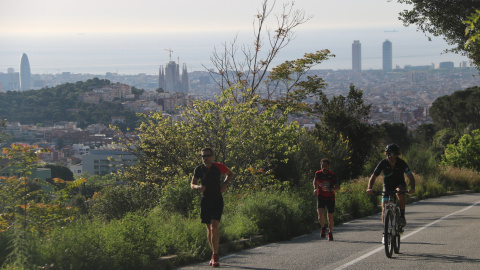 Pla general d'un ciclista i dos corredors pujant pel carrer Manuel Arnús, just abans d'arribar a la carretera de les Aigües, al Parc de Collserola de Barcelona. Imatge del 2 de maig de 2020. (Horitzontal)