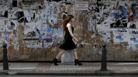 Una mujer con mascarilla camina este jueves por una céntrica calle de A Coruña, frente a un muro lleno de carteles con actividades culturales,en la jornada en la que ha entrado en vigor llevar este tipo de protección en espacios públicos, tanto al air