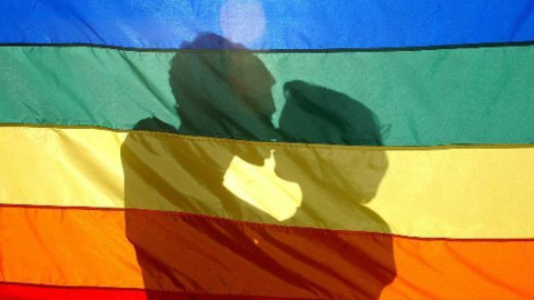 Una pareja se besa tras la bandera del arcoíris, símbolo del movimiento LGTBI.