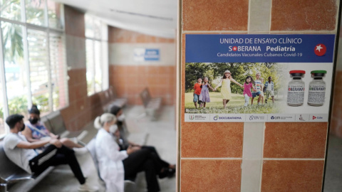 Imagen de archivo de varias personas sentadas cerca de un cartel que anuncia los ensayos clínicos de la vacuna Soberana 02, en una clínica sanitaria en La Habana. - REUTERS