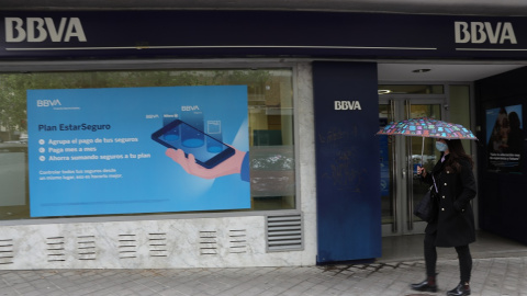 Una mujer con paraguas pasa por delante de una oficina de BBVA en Madrid. E.P./Isabel Infantes