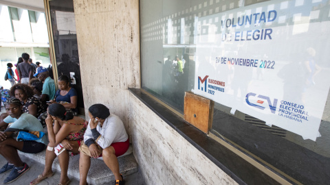 Varias personas permanecen sentadas junto a un cartel de las elecciones locales del 27 de noviembre.