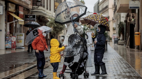 Una mujer con tres menores se protegen de la lluvia con paraguas y chubasqueros en una céntrica calle de València.