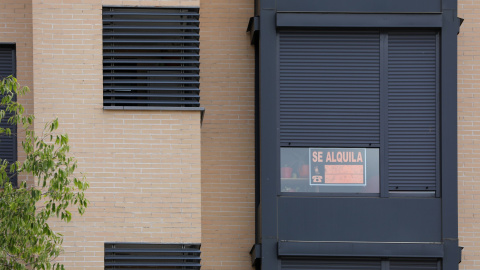 En la fachada de un edificio se ve un cartel de 'Se Alquila' bajo la persiana de uno de los pisos.