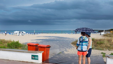 Dos mujeres se resguardan de la lluvia bajo un paraguas en una playa de Menorca | EFE/Archivo