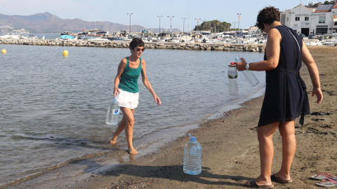 Dues dones omplen ampolles i garrafes del mar a la platja del Port de la Selva