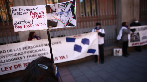 Una mujer con un cártel en el que se lee: `Por dignidad y derechos para los vivos, Justicia para los fallecidos´ durante una concentración de familiares de usuarios de residencias frente a la Consejería de Políticas Sociales "por una gestión públic