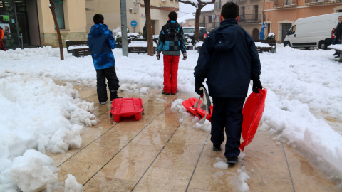 Trens nens amb trineus creuen la plaça plena de neu a Horta de Sant Joan, durant el temporal del febrer del 2023