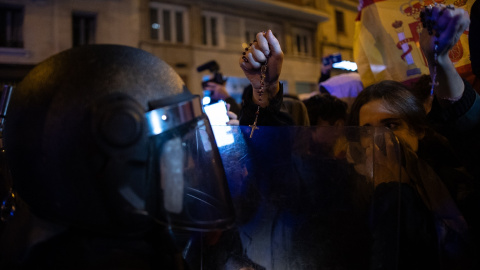 Un manifestante embozado con dos rosarios en las manos se enfrenta a un agente de Policía, durante una protesta contra la amnistía frente a la sede del PSOE.