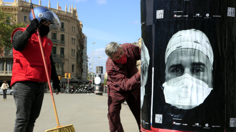 Cartells amb cares de professionals sanitaris amb mascareta en una columna publicitària de plaça de Catalunya de Barcelona, en el projecte 'Behind the mask', que busca el reconeixement col·lectiu davant de la covid-19. 8 d'abril del 2021.