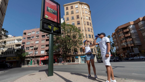 Dos jóvenes beben agua junto a un termómetro que marca 43 grados centígrados, en la Plaza del Royo de Murcia.