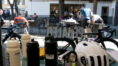 En primer terme, cascos i bidonets i, al fons, uns ciclistes en una terrassa de Girona