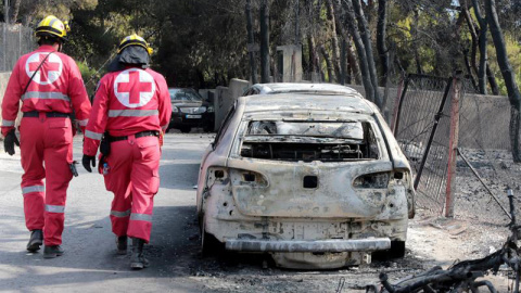 Miembros de la Cruz Roja caminan junto a un vehículo calcinado en una zona afectada por los incendios en Argyra Akti, en Mati. (PANTELIS SAITAS | EFE)