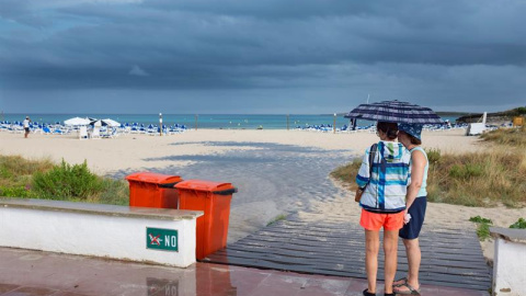 Dos mujeres se resguardan de la lluvia bajo un paraguas en la playa de Punta Prima, en el municipio de Sant Lluís (Menorca), en una jornada en la que la Agencia Estatal de Meteorología (Aemet) ha activado la alerta amarilla en Baleares por posibles prec