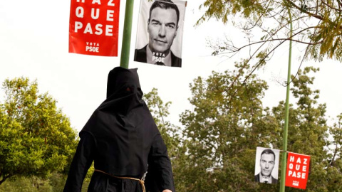 Un penitente de la Hermandad de La Sed camina por un acella de Sevilla con un cartel electoral del PSOE. (MARCELO DEL POZO | EFE)