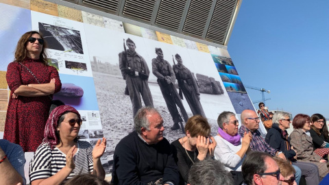 Assistents a la inauguració del mural en memòria dels afusellats al Camp de la Bota, davant de fotografia de soldats de l'exèrcit en pràctiques de tir en aquell mateix lloc. PÚBLIC