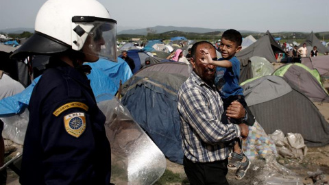 Un agente antidisturbios junto a un padre y su hijo en uno de los campamentos de refugiados improvisados en Idomeni, Grecia. - EFE