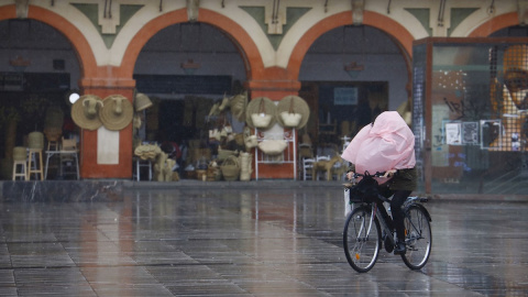 Una mujer en bicicleta a la que el viento le ha tapado la cara totalmente con el chubasquero mientras pasa por la Plaza de la Corredera de Córdoba