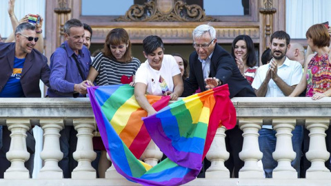 El alcalde de Valencia, Joan Ribó (centro), despliega desde el balcón del Ayuntamiento la bandera arco iris con motivo del Día Internacional del colectivo LGTB. /EFE