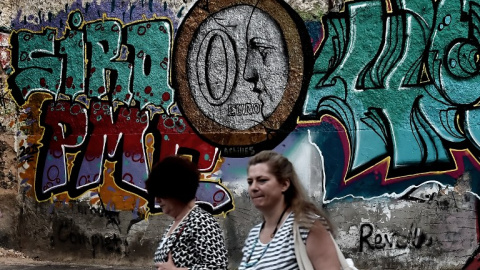 Dos mujeres caminan frente a un muro con un grafitti en el que aparece una moneda de euro con un 0 en su interior. AFP