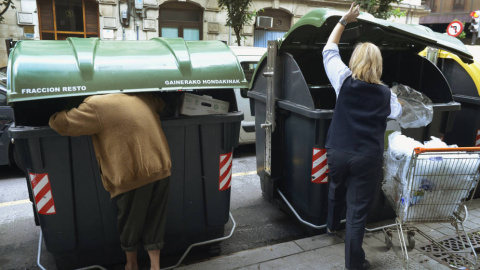 Una persona busca comida en un contenedor de basura de Bilbao, mientras una trabajadora de un supermercado echa bolsas a la basura. REUTERS