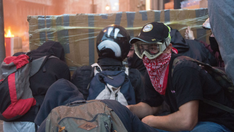 Varios jóvenes encapuchados se protegen de las balas de goma con un parapeto de cartón y cinta de embalaje, durante los disturbios de Barcelona. GUILLEM SANS