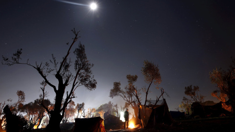 Un refugiado se calienta junto al fuego en un improvisado campamento cerca del centro de registro en la isla griega de Lesbos.- REUTERS / Yannis Behrakis