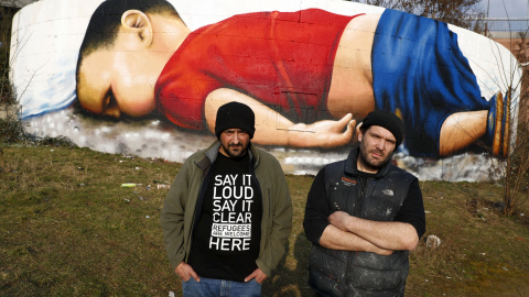 Artistas Justus Becker (R) y Oguz Sen se paran frente a una gran obra de arte pintada del niño sirio Aylan Kurdi, después de terminar una sesión de pintura de tres días en una pared en las orillas del río Main, cerca de la sede del Banco Ce