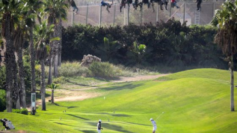 Una docena de subsaharianos permanece encaramada a la valla de Melilla, mientras dos personas juegan al golg. José Palazón tituló esta imagen como ‘Paisajes de desolación’.