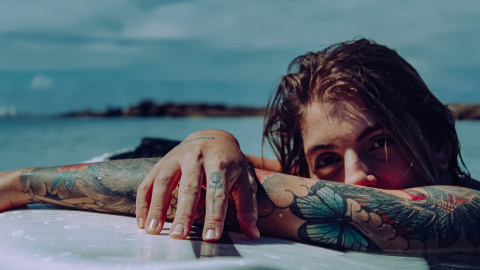 Una mujer tatuada descansa sobre una tabla de surf en la playa. Foto: Pexels