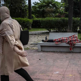 Una mujer observa a Mohamed, un menor marroquí de 17 años que duerme en el banco de los Jardines de Argentina de Ceuta tras entrar a nado a la ciudad a principios de esta semana.- JAIRO VARGAS