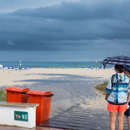 Dos mujeres se resguardan de la lluvia bajo un paraguas en una playa de Menorca | EFE/Archivo