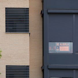 La fachada de un edificio donde se ve un cartel de 'Se Alquila' bajo la persiana de uno de los pisos.