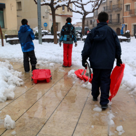 Trens nens amb trineus creuen la plaça plena de neu a Horta de Sant Joan, durant el temporal del febrer del 2023