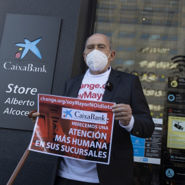 Carlos San Juan, el hombre de 78 años que ha recogido más de 600.000 apoyos con su campaña 'Soy Mayor, No Idiota', posa delante de una sucursal bancaria con un cartel que simula la entrega de sus firmas a los bancos, tras registrarlas en el Ministerio 